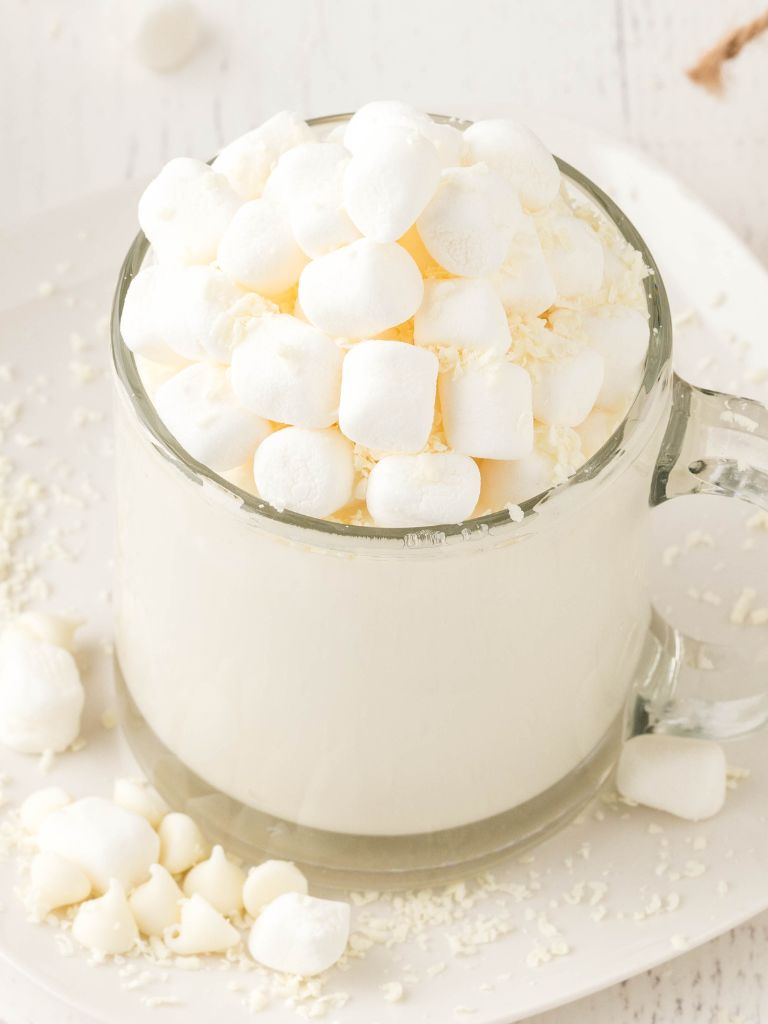 Side shot of hot chocolate with marshmallows in a clear mug sitting on a white plate. 