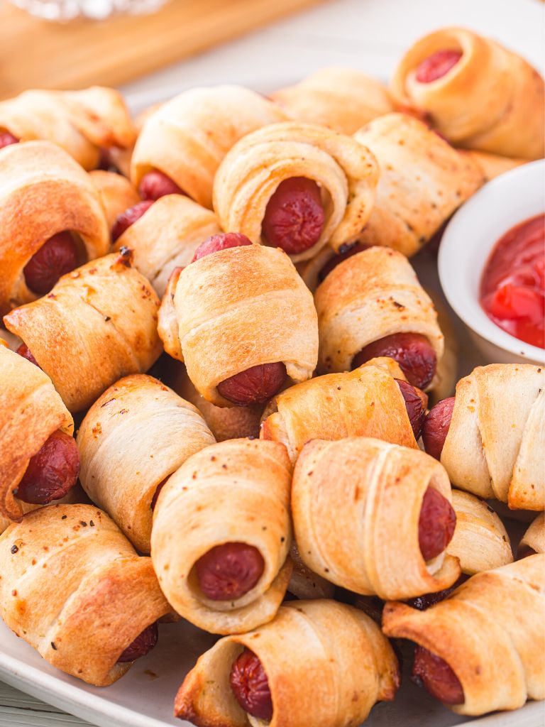A plate of pigs in a blanket with a small bowl of ketchup next to them on the plate. 