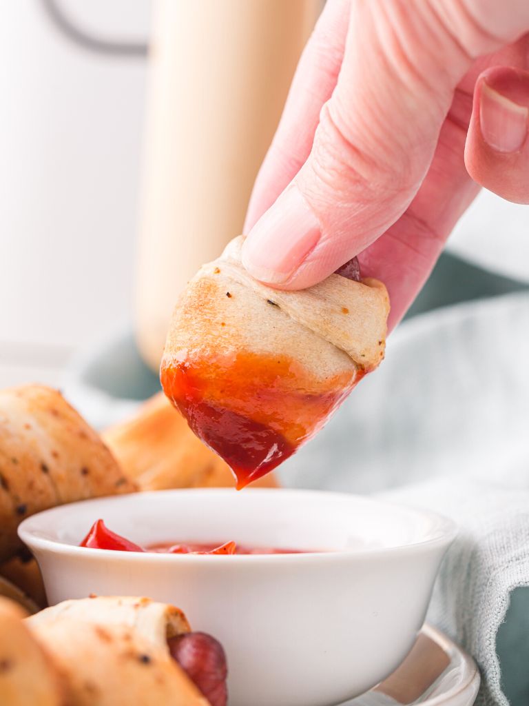 A hand dipping a pig in a blanket in some ketchup. 