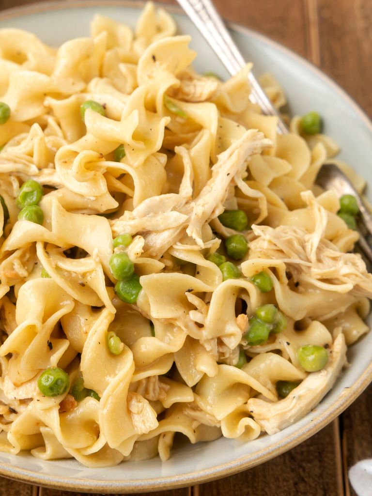 Egg noodles and chicken in a creamy mixture laying on a white plate against a wooden background table. 