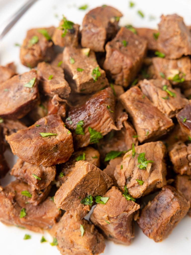 A pile of steak cut into cubes sitting atop a white plate. 