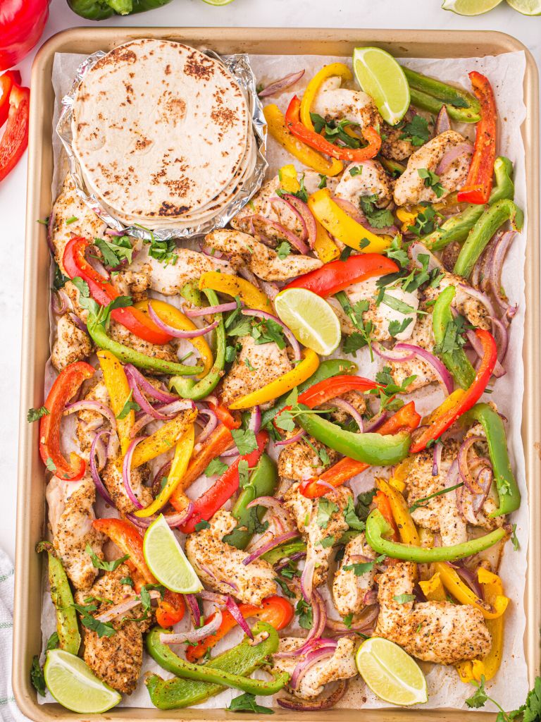 Overhead photo of a sheet pan with chicken and bell peppers on it and a stack of tortillas. 