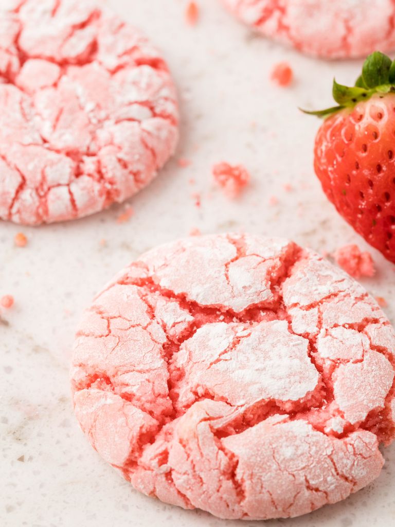 Close up picture of a cookie with more in the background and a fresh strawberry near. 