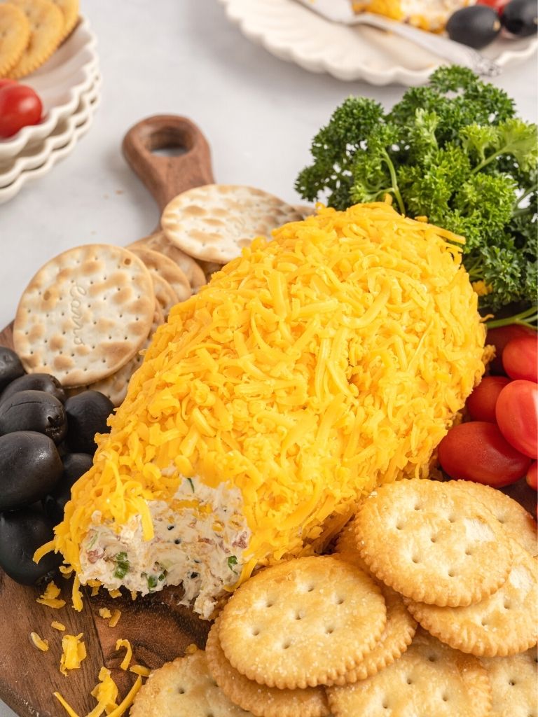 A cheeseball with cheese and parsley on the outside on top of a wooden cutting board. 