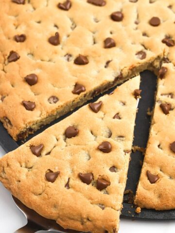 A cookie pizza with a piece cut out of it on a server spoon.