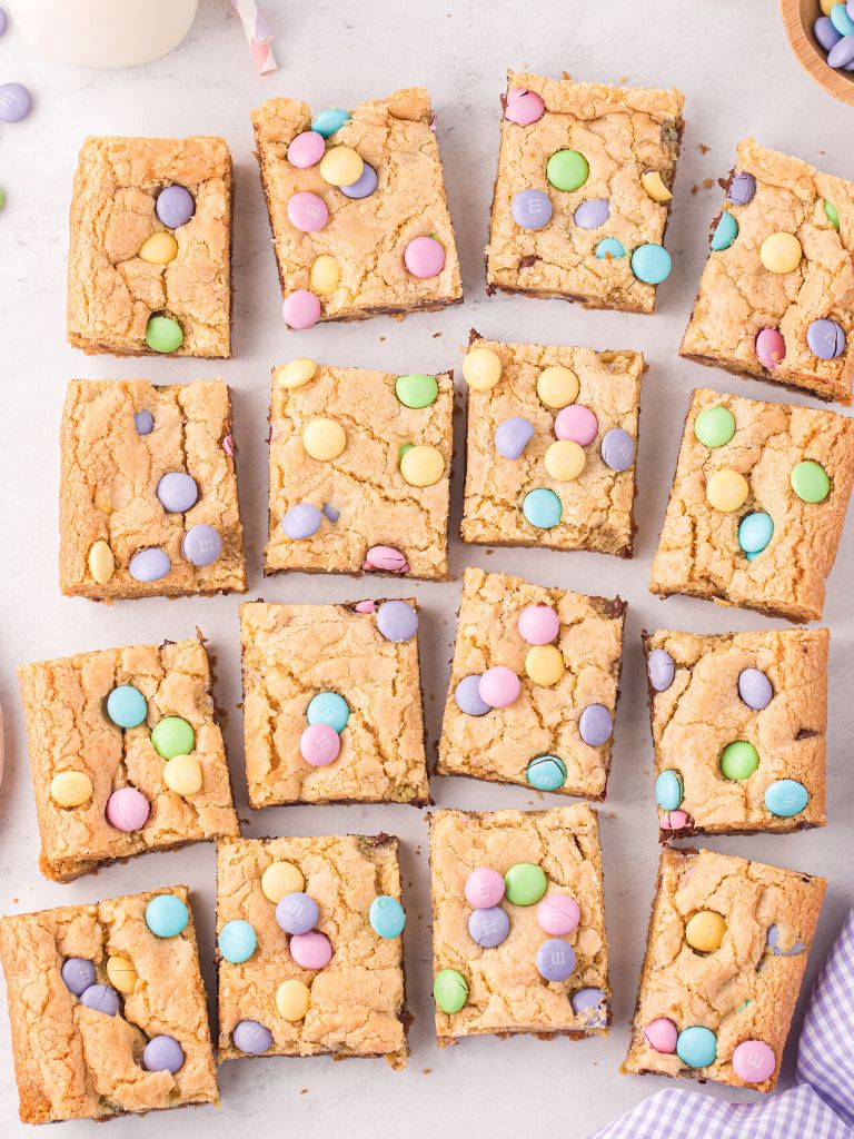 Overhead shot of squares of cookie bars with m&m on top and some around it, a cloth, and a cup of milk. 