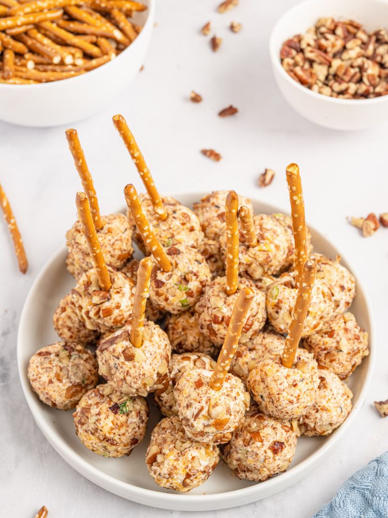 A plate of appetizers with pretzels and cream cheese balls with a bowl of ingredients in the background. 