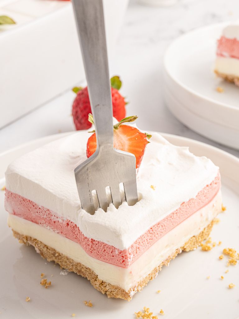 A fork going into a slice of this dessert with layers of strawberry and cheesecake. 