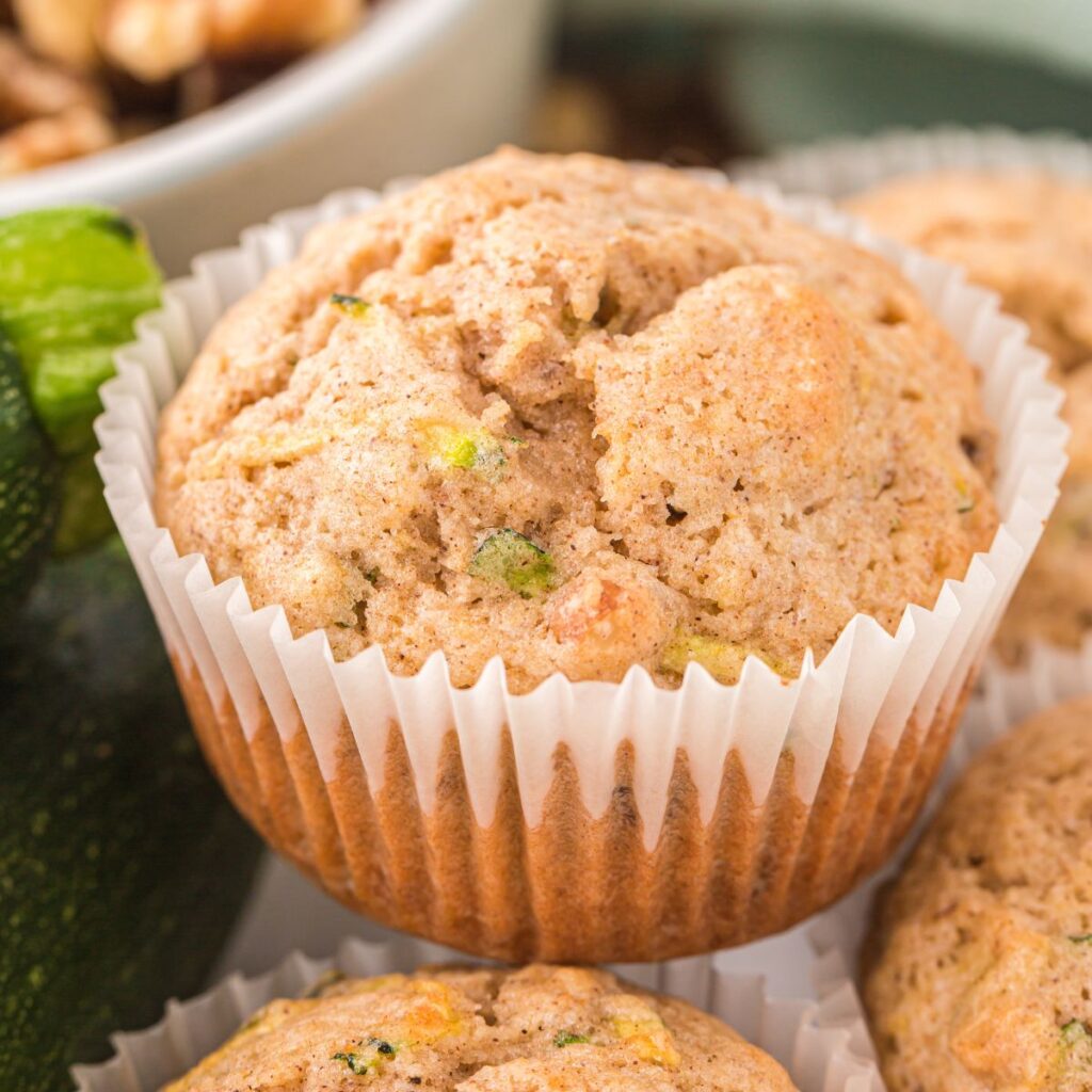 A muffin up close inside a wrapper with a fresh zucchini next to it.