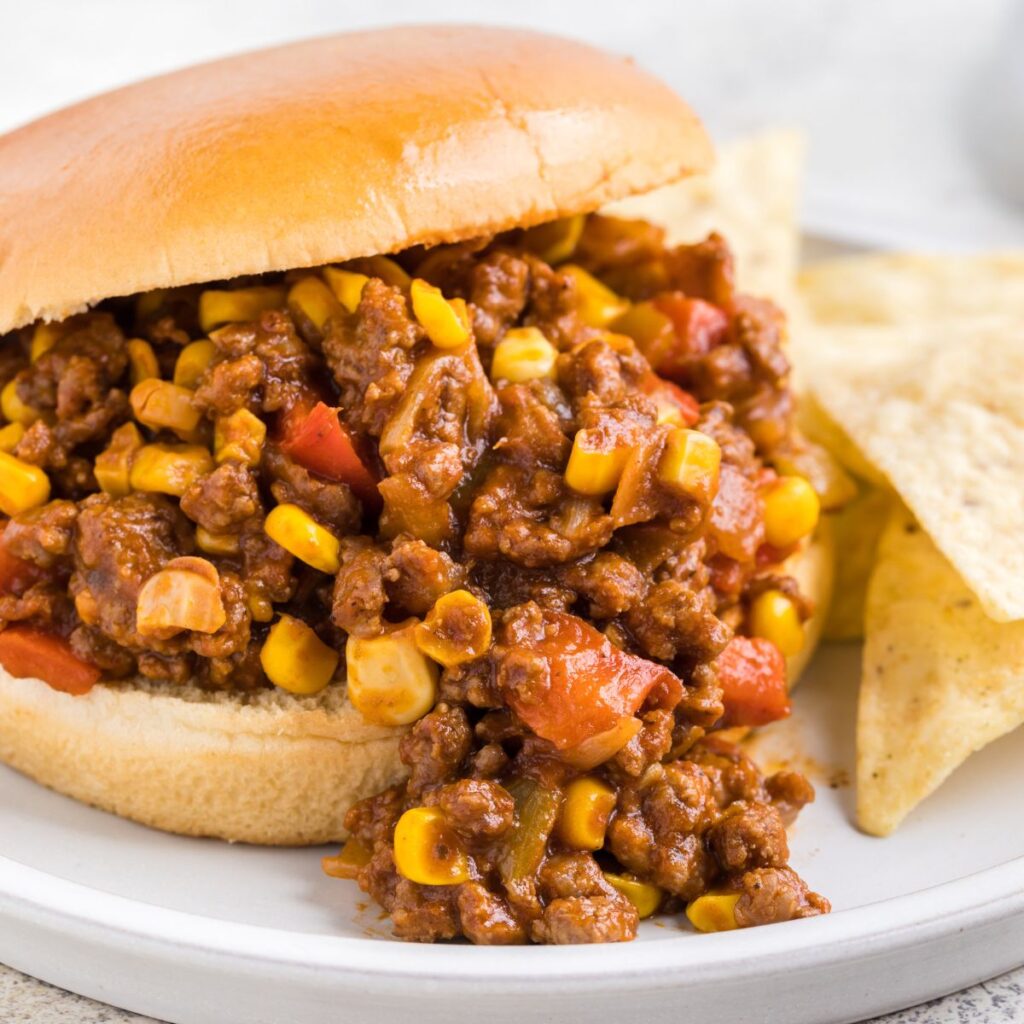 A photo of a sloppy Joe on a white plate next to tortilla chips. 