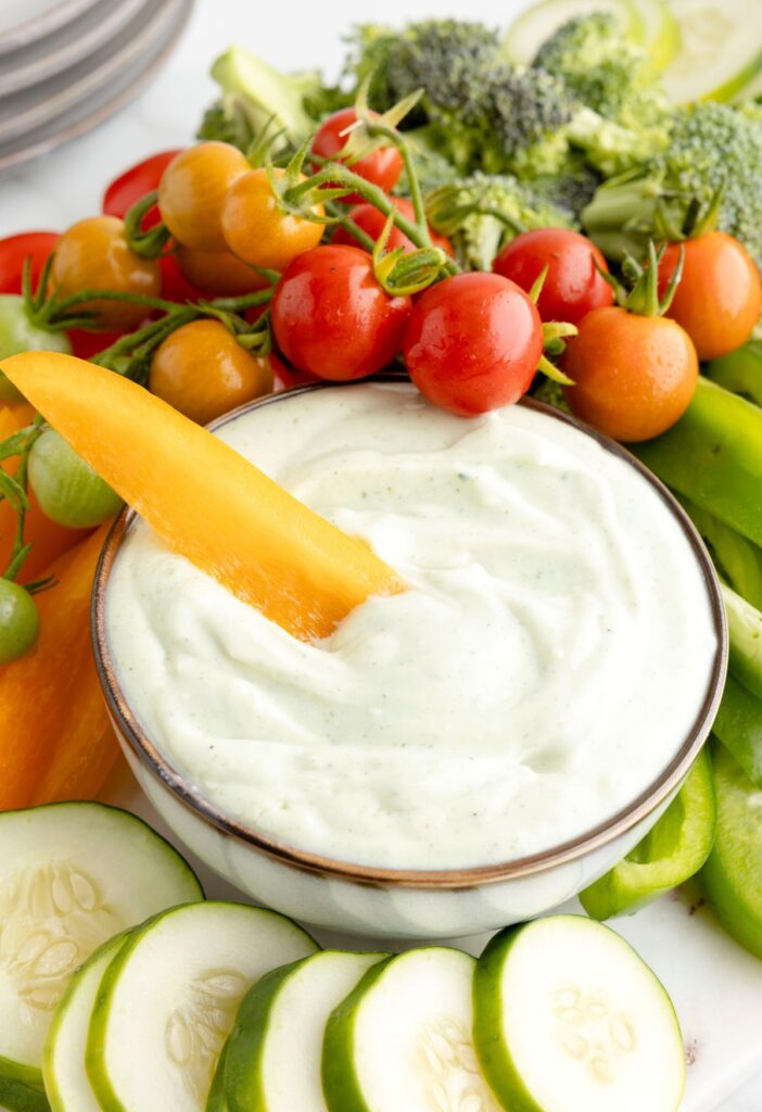 A bowl of dressing with veggies around it and a bell pepper stick in the ranch. 