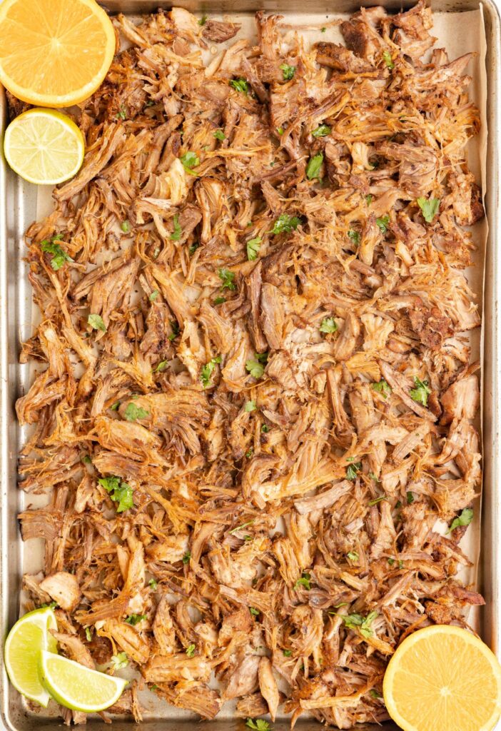 A sheet pan of carnitas made with pork with fresh citrus on the edges of the pan. 