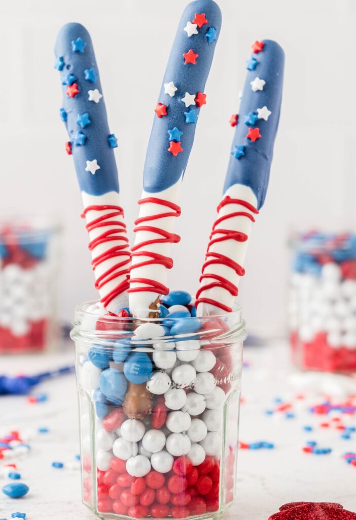 A cup with red white blue candies in it and the decorated pretzel sticks. 