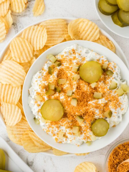 A bowl of dip garnished with pickles and chips.