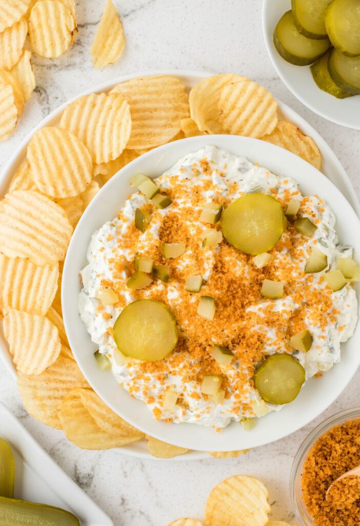 A bowl of dip garnished with pickles and chips. 