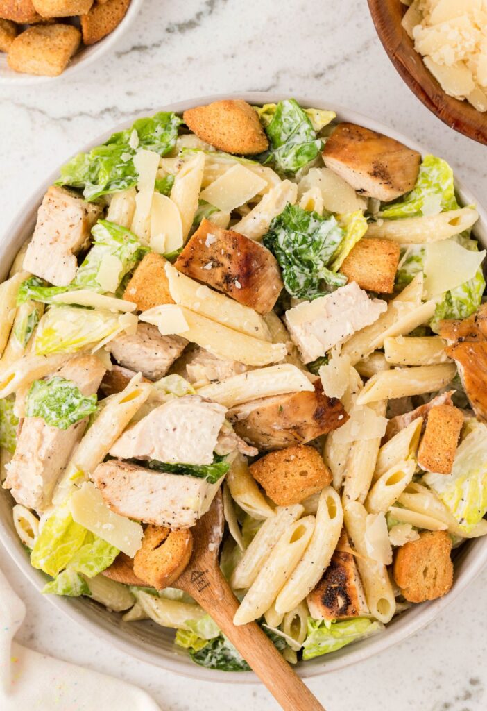A serving bowl of pasta salad with wooden spoon and croutons on the side. 