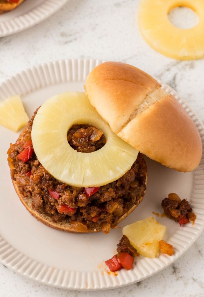 Sloppy Joe meat on a bun with a pineapple on top, sitting on a white plate. 