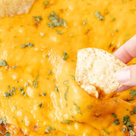 A photo of cheesy bean dip with a finger holding a chip for dipping.