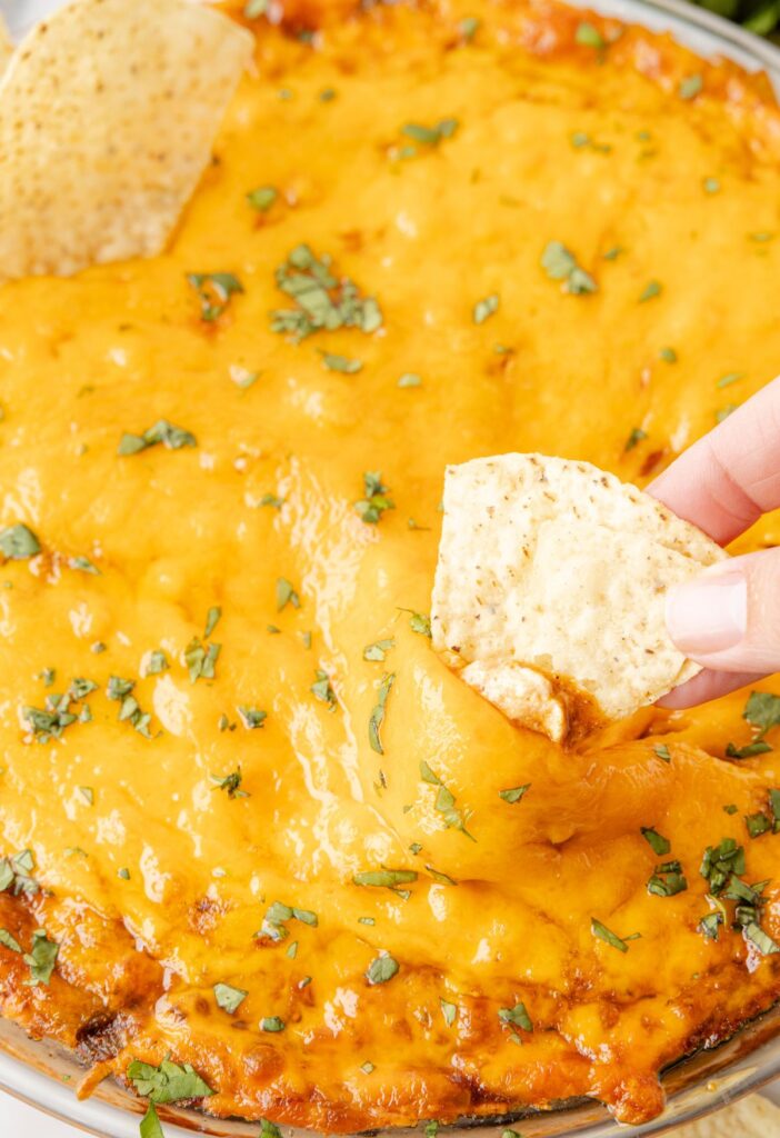 A photo of cheesy bean dip with a finger holding a chip for dipping. 