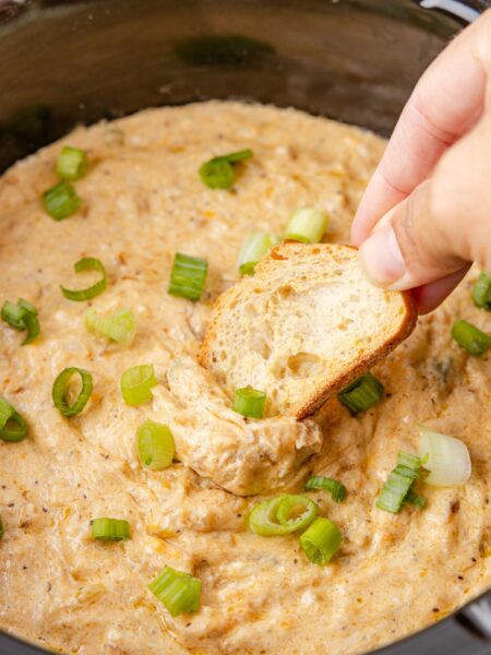 A crock pot with dip inside of it and hand holding a piece of bread over it