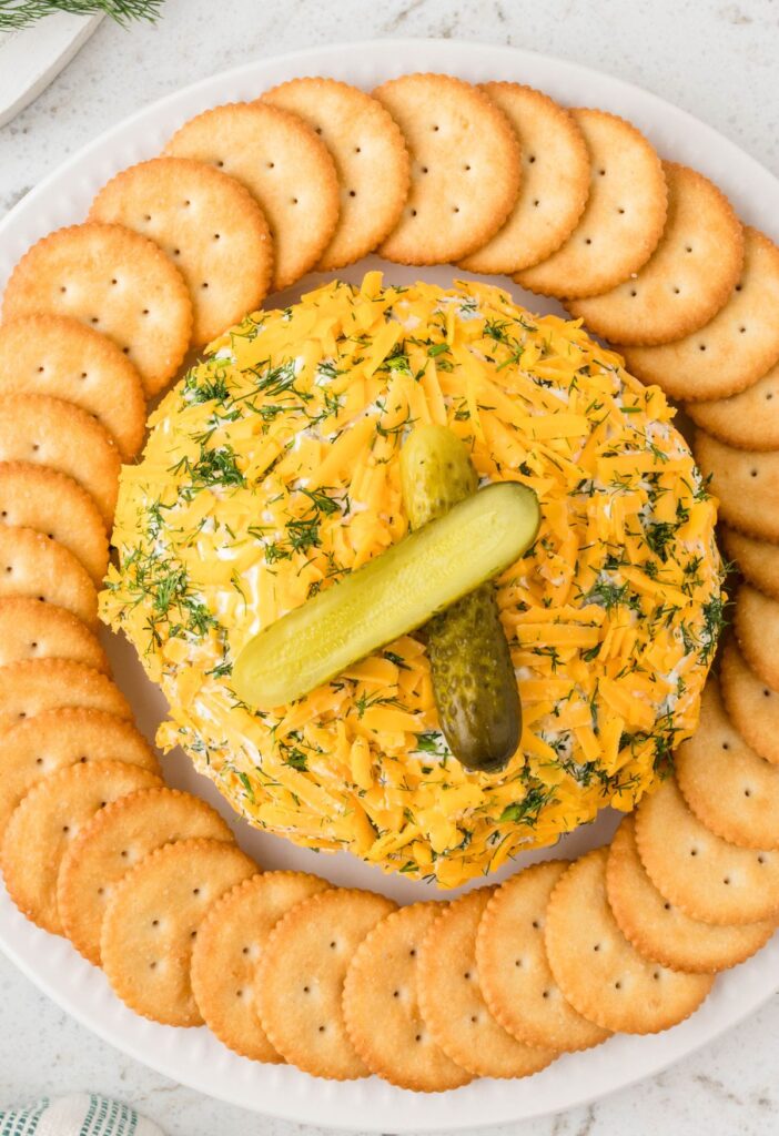 Top view of the appetizer cheeseball with a pickle on top of it and crackers around it. 