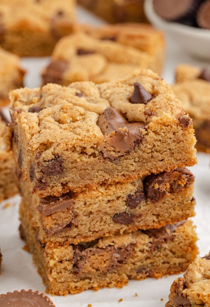 A stack of cookie bars with a bowl of peanut butter and Reeses peanut butter cup behind them. 