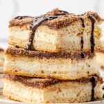 A stack of cheesecake bars with churro ingredients and chocolate sauce over top
