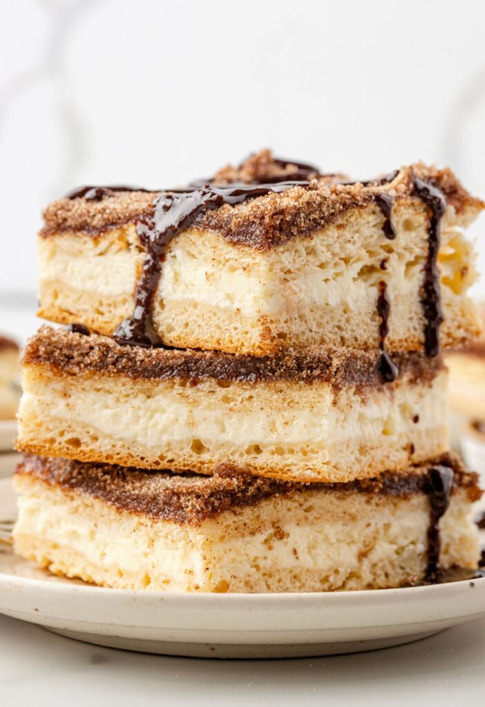 A stack of cheesecake bars with churro ingredients and chocolate sauce over top