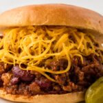A sloppy Joe on a white plate with chili cheese mixture and cheese.
