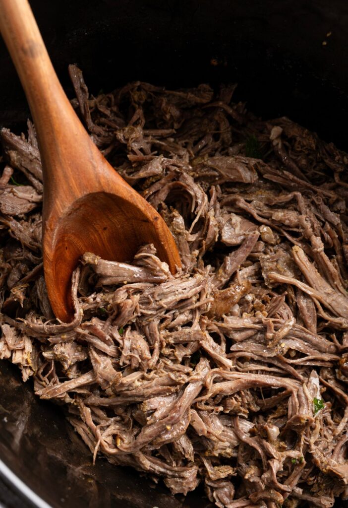 Inside of the crockpot with the shredded beef and a wooden serving spoon in it. 