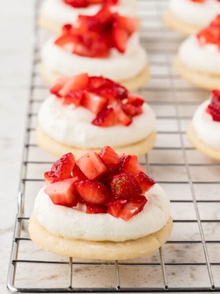 Sugar cookies with whipped cream and chopped strawberries sitting on a cooling rack.