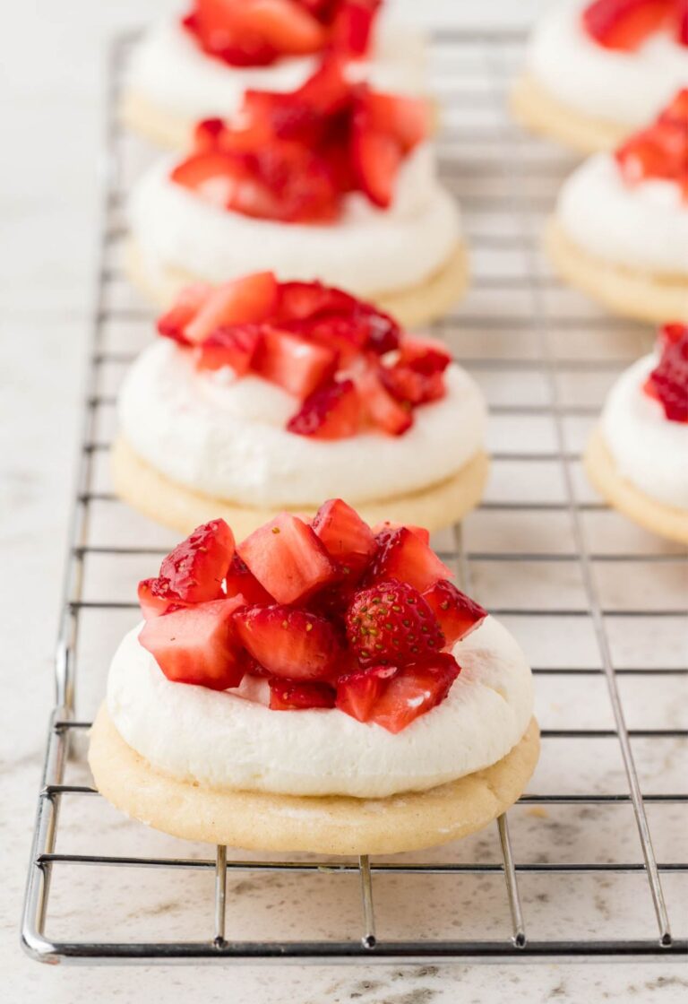 Sugar cookies with whipped cream and chopped strawberries sitting on a cooling rack.