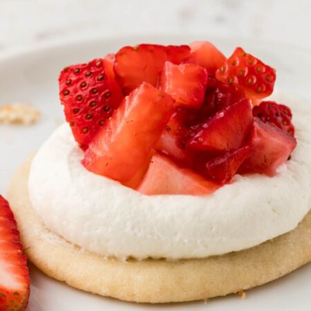 A strawberry shortcake cookie sitting on a white plate with a whole strawberry next to it.