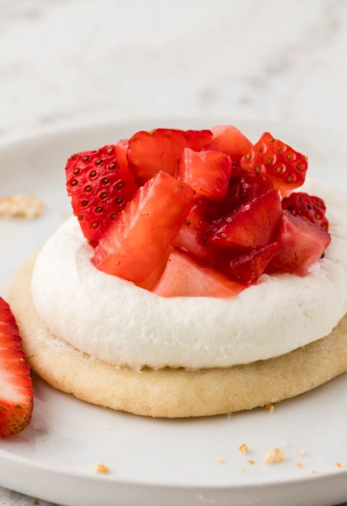A strawberry shortcake cookie sitting on a white plate with a whole strawberry next to it. 