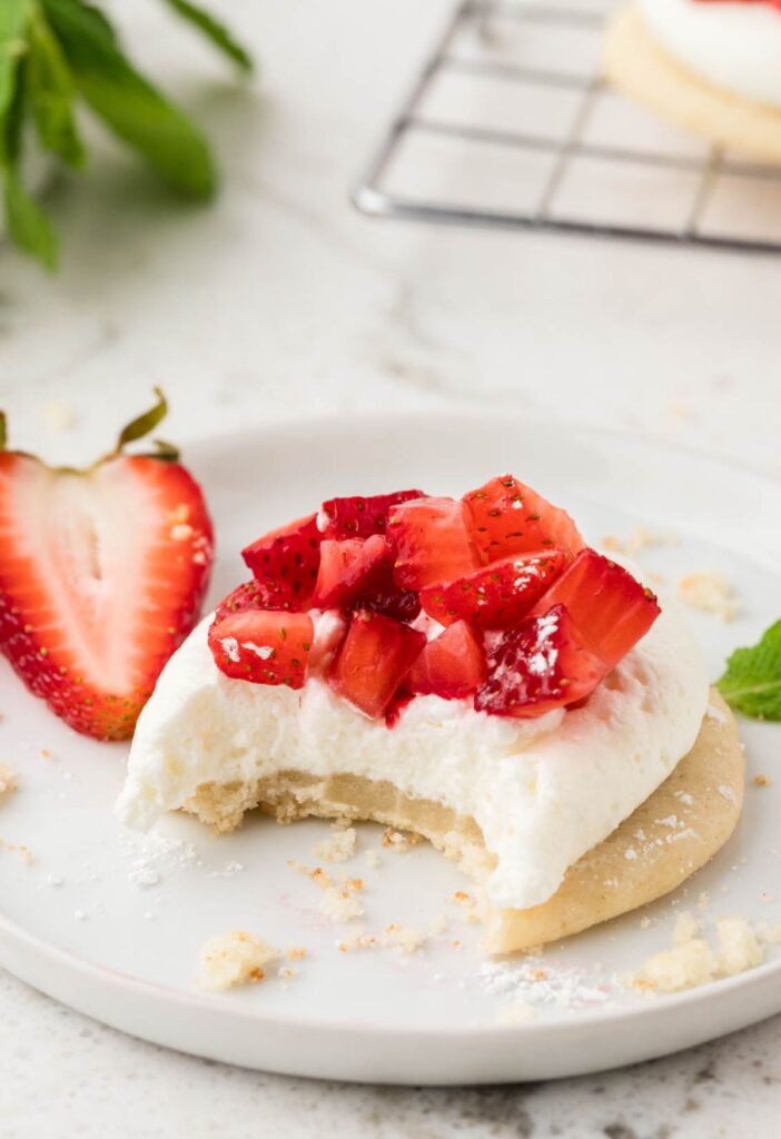 A cookie with whipped cream and strawberries on top of it, sitting on a white plate, with a bite taken out of it. 