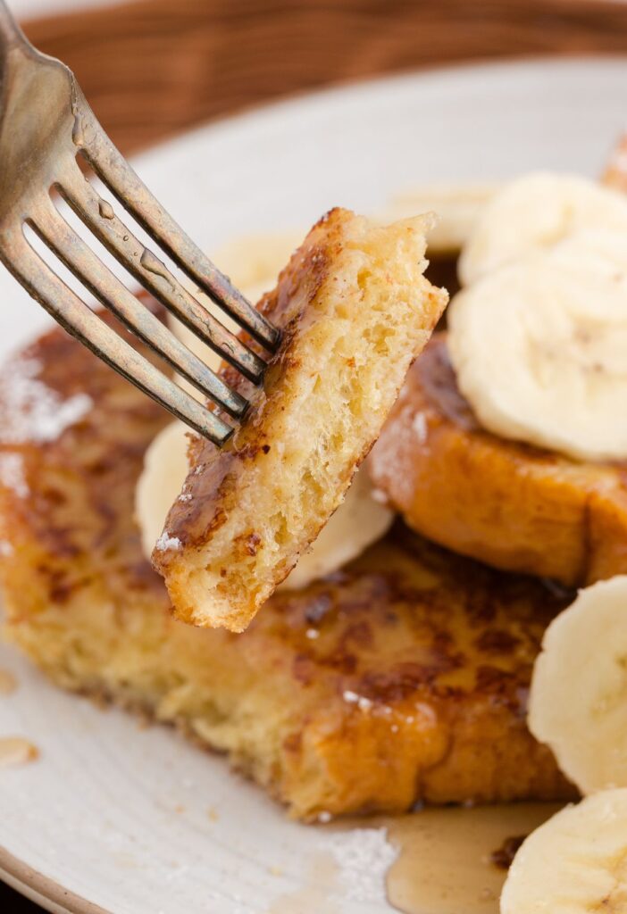 A fork with a bite of French toast on it above the serving plate of the French toast slices. 