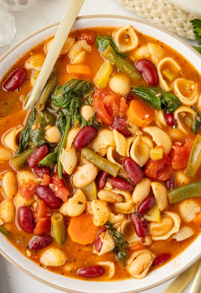 A bowl of veggie loaded minestrone soup with a gold spoon.