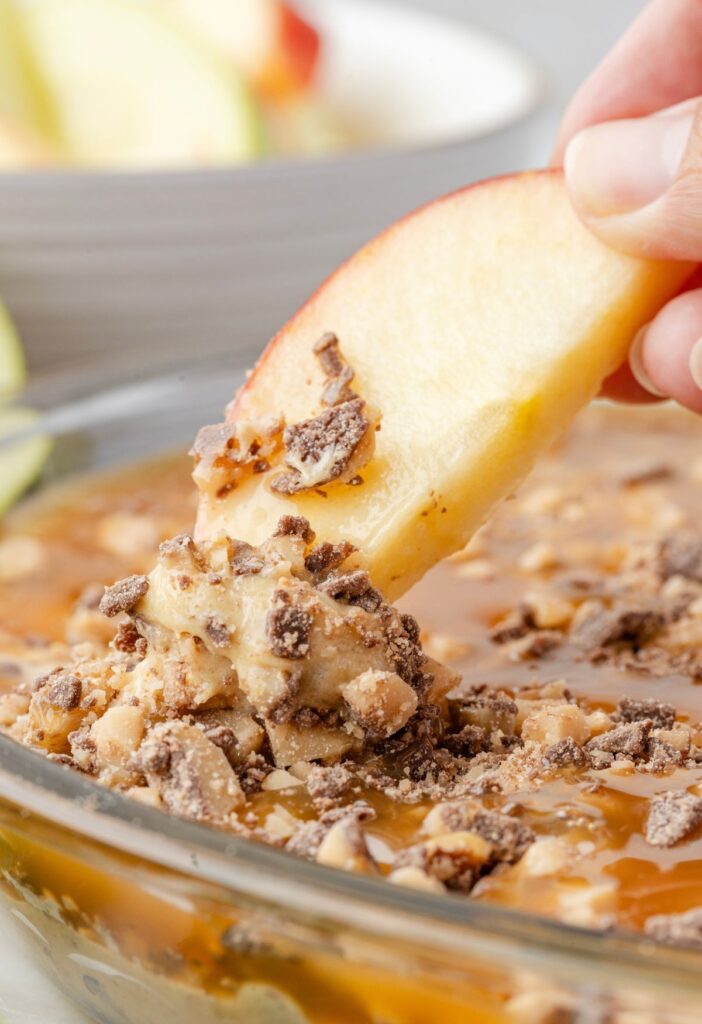 A apple slice being dipped into the caramel apple fruit dip, with fingers in the pic holding the apple slice 