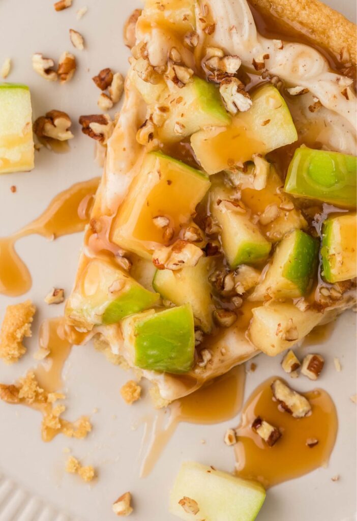 A half eaten piece if caramel apple pizza on a white plate with caramel sauce