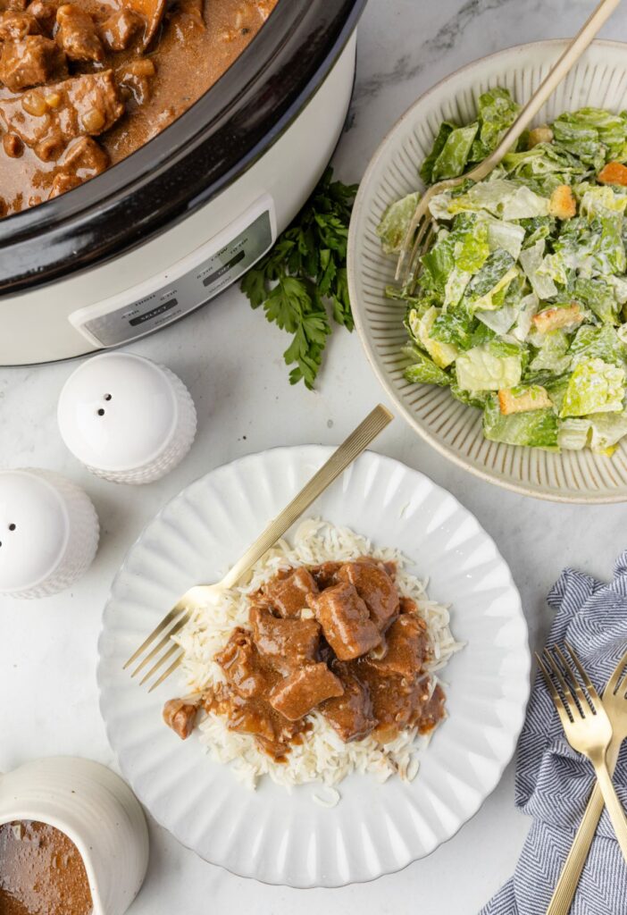 Overhead pic of the slow cooker with the beef tips inside, a serving plate of some, and a green salad. 