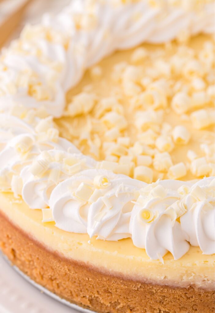 A whole cheesecake, side view, with white chocolate curls on top and whipped cream
