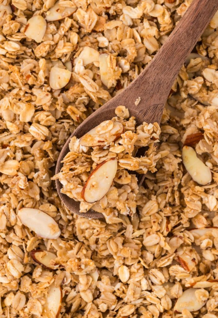 A cookie sheet with oven baked granola on it and a wooden serving spoon scooping some up.