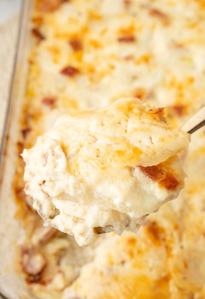 A serving spoon with some potatoes on it above the casserole dish. 