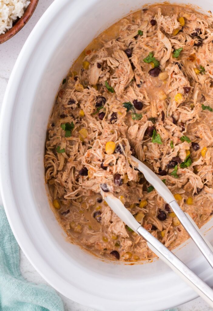 Tongs inside the slow cooker scooping up some shredded fiesta chicken