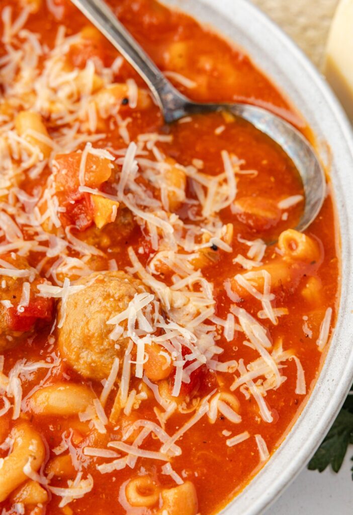 Serving bowl of meatball soup topped with parmesan cheese and a spoon in it. 