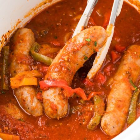 A white slow cooker with sausages in tomato sauce and tongs.