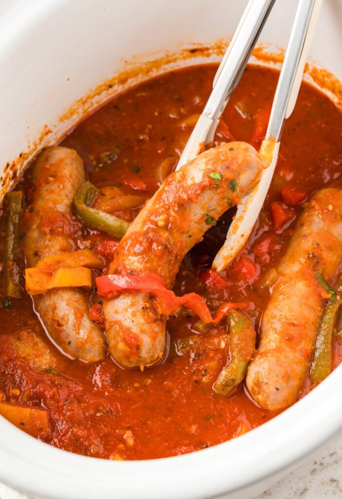 A white slow cooker with sausages in tomato sauce and tongs. 