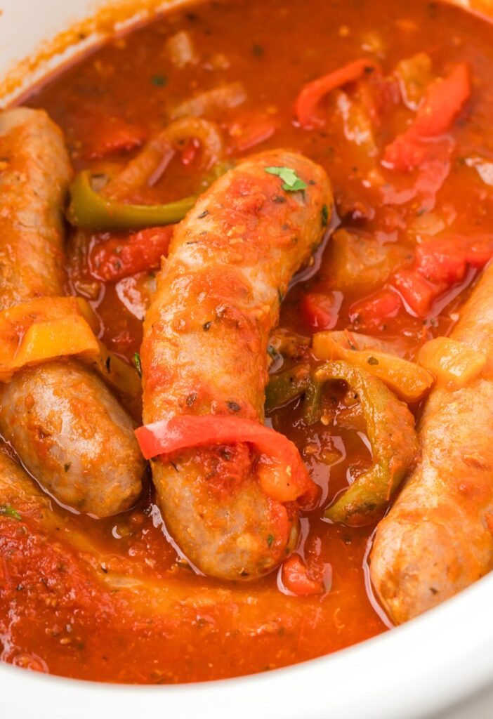 Inside of the white slow cooker showing the sausages and peppers and onions. 