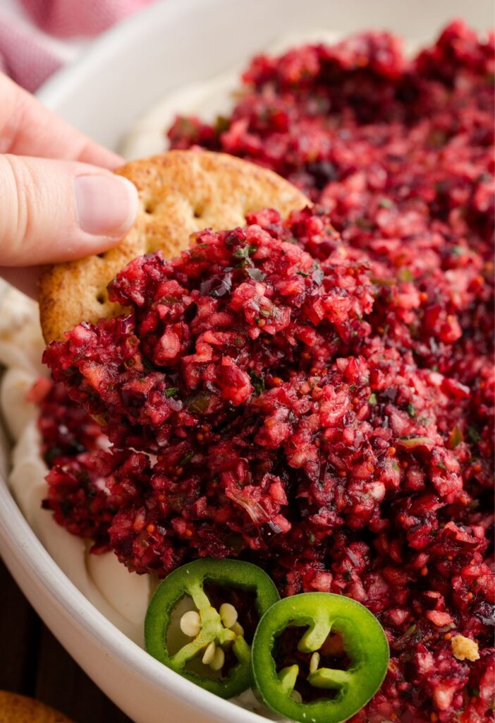 A white bowl of cream cheese dip with a cranberry topping, a hand holding a cracker with some dip on it.