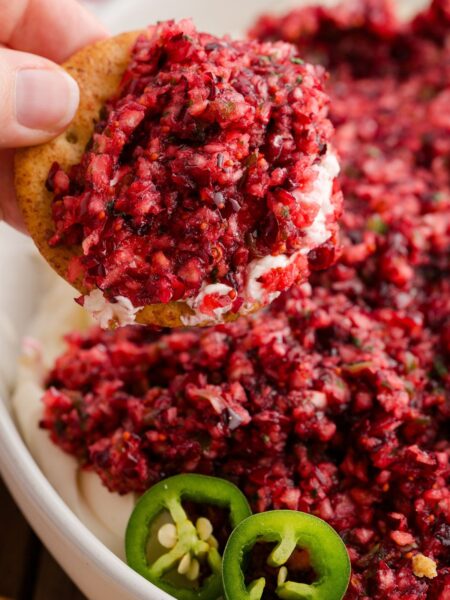 Cream cheese dip with a cranberry and jalapeno topping, a hand holding a cracker ready to dip.